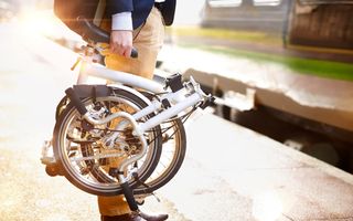 Een man met een opgevouwde vouwfiets in zijn rechterhand wacht op een perron op de trein.
