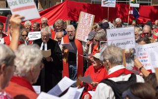 Een groep demonstranten, onder wie een man in een zwarte toga, houdt borden met leuzen in de lucht.