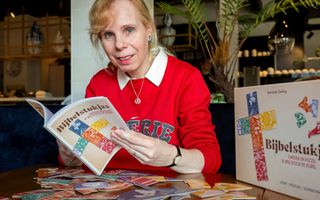 Een oudere vrouw met een rode trui aan een tafeltje met allemaal puzzelstukjes erop. Ze houdt het boekje ”Bijbelstukjes” omhoog; rechts de doos waarin een en ander zit.