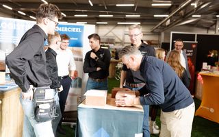 Zes mannen staan om een tafel waarop rijtjes bakstenen liggen. De man rechts houdt zo’n rijtje stenen tussen zijn beide handen. 