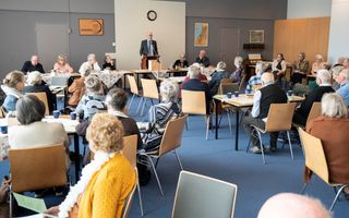 Een man met bril, in pak, staat voor een zaal vol oudere mensen die aan tafeltjes zitten.