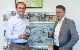 Twee mannen houden samen een landkaart vast. Ze dragen allebei een overhemd. De man rechts draagt ook een grijs colbert.