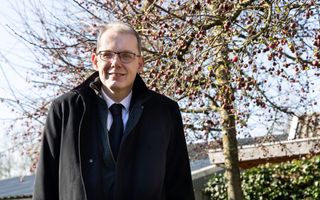 Een predikant met bril in een zwart pak met stropdas en een zwarte overjas staat in een tuin voor een boom.