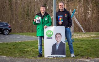 Een jongeman en een man van middelbare leeftijd staan bij een lantaarnpaal, met een flyer van hun partij in de hand. Om de lantaarnpaal zit een verkiezingsposter waarop een foto van de jongeman staat.