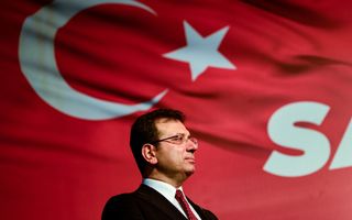 Een man met zwart haar, vierkante bril, in zwart pak en rode das staat voor een grote Turkse vlag. De vlag is rood met een halve maan en een ster. 