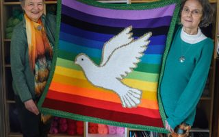 Twee lachende vrouwen houden veelkleurig kleed met daarop witte duif vast. 