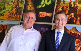 Een wat oudere man met wit overhemd staat links, een jongeman met donkerblauw pak en stropdas staat rechts. Achter hen een aantal schilderijen met Bijbelse afbeeldingen, zoals Adam in het paradijs, de slang in de hof en andere taferelen.