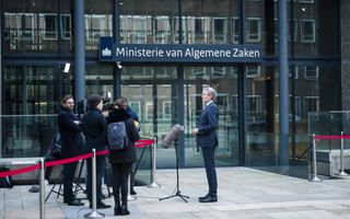 Voor een groot gebouw op de achtergrond staat een man in pak enkele mensen van de pers te woord.