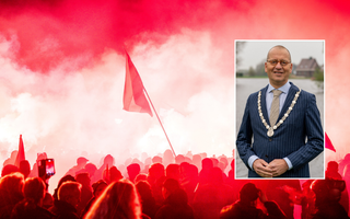 Burgemeester met ambtsketting om en in blauw krijtstreeppak en bril op voor een rivier. Op de achtergrond een foto van een protest met veel rode rook.