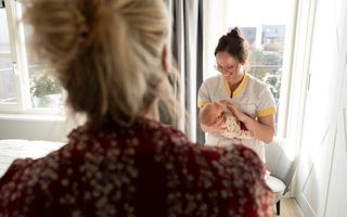 Op de voorgrond vaag een vrouw met blond haar, op de achtergrond een kraamverzorgende met baby. 