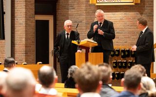 Drie mannen in een zwart pak staan voor in een kerkzaal, tegenover mensen die in de banken zitten. De middelste man staat achter een katheder.
