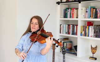 Vrouw in blauw-witte blouse speelt viool voor een witte boekenkast. 