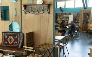 Een wand met een lege kapstok. Rechts zitten twee mannen aan een tafel.