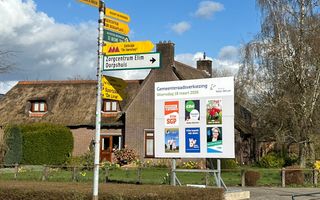 Een verkiezingsbord. Op de achtergrond een woning met een rieten dak. 