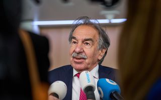 Oudere man met snor in pak staat voor drie microfoons.