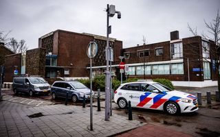 Voor een groot gebouw staan een busje en een auto geparkeerd. Daarvoor rijdt een politieauto. Aan een paal hangt een beveiligingscamera.