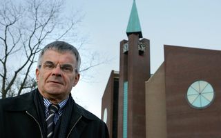 Een man met stropdas en bril staat voor een kerkgebouw met een groene toren.