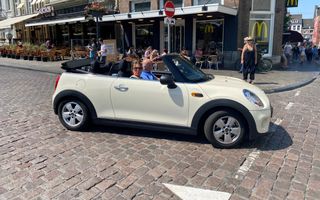 Een witte cabriolet rijdt door het centrum van Maastricht. Op de achtergrond een fastfoodrestaurant en wat terrasjes. 