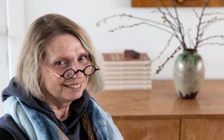 Foto van een oudere vrouw met kort grijs haar en een ronde bril. Ze draagt een blauwe shawl. Op de achtergrond een lage bruine kast met een stapeltje boeken en een vaas met takken.