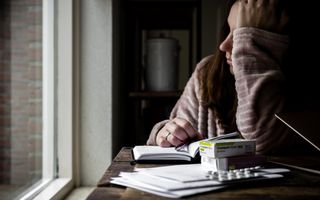 Vrouw in een badjas steunt met haar hand tegen haar hoofd. Voor haar liggen papieren en medicijnen.