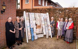 Acht dames naast een oud droogrek waar allemaal borduurwerk overheen hangt. 