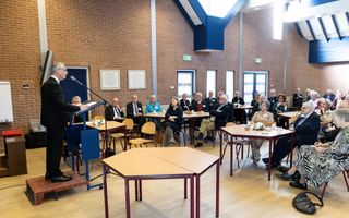Een man in donker pak spreekt de zaal met oudere mannen en vrouwen toe. Ze zitten aan tafeltjes.