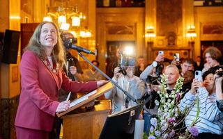 Vrouw in roze mantelpakje tuurt naar een beeldscherm. Om haar heen staan tal van mensen met camera’s en smartphones in de aanslag.