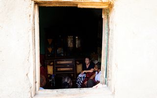 Inkijkje van buiten naar binnen genomen. Twee vrouwen op een bank in gesprek met een gezin.  