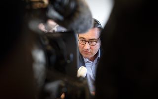 Camera’s zijn gericht op een man met pak en bril, die in een microfoon praat.