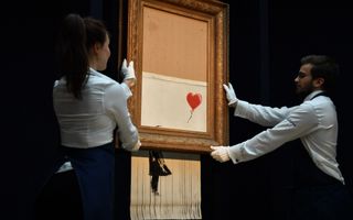 Twee mensen in witte blouses poseren met een kunstwerk dat half uit een lijst hangt en aan de onderkant is gerafeld. 