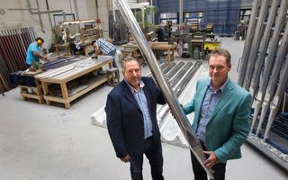 Twee mannen in pak staan in een bedrijfshal. Achter hen liggen en staan orgelpijpen. Samen houden ze een grote orgelpijp vast.