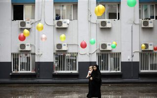 Vrouw in het zwart gekleed en met hoofddoek met een kind op de arm. Achter haar een wit gebouw. Gekleurde balonnen hangen op het plein. 