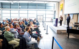 Een groep mensen in een zaal luistert naar een spreker.