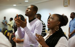 Een tiental mensen, met een donkere huid, staat met gesloten ogen en bidt of klapt in de handen.