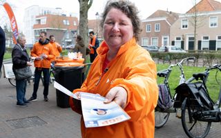 Vrouw met bruin haar en oranje jas deelt flyers uit op straat. 