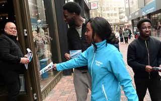 Een vrouw in een blauwe ChristenUniejas deelt een flyer uit aan een man in de deuropening van een winkel. 