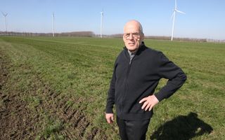 Een ernstig kijkende, kale man in zwarte kleding staat in een groen weiland. Op de achtergrond zijn windmolens te zien.