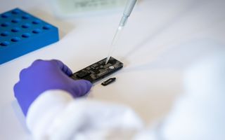 Een hand gestoken in een blauwe handschoen houdt een apparaatje en een pipet vast.