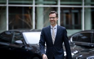 Een man met baard in pak met op de achtergrond een aantal donkere auto’s.