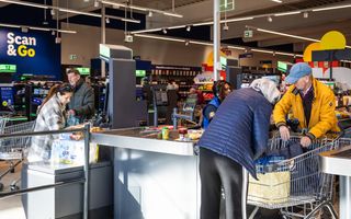 En man en een vrouw legen hun boodschappenkarretje bij een kassa in een supermarkt. Op de achtergrond rekent een andere vrouw haar boodschappen af bij een zelfscankassa.