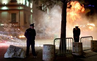 Twee politieagenten staan naast betonblokken en dranghekken. Op de achtergrond is veel vuurwerk, vuur en rook te zien.