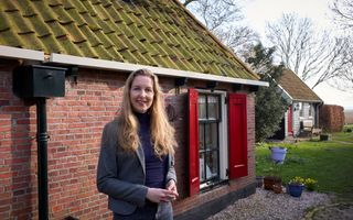 Een vrouw met lang, blond haar en een grijs jasje aan bij een oud huisje met rode luiken.
