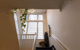 Bovenaan een trap staat een witte kaars op een zwarte standaard. Voor de standaard staan oranje bloemen. 