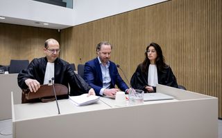 Links een man in toga, middenin een man in blauw pak, rechts een vrouw in toga. Ze zitten achter een witte desk met microfoons.