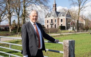 Een man in kostuum met oranje stropdas in het zonnetje voor een groot landhuis.
