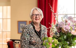 Oudere vrouw in veelkleurig jasje. Rechtsvoor een bos bloemen, op de achtergrond een rood gordijn. 