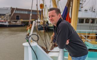 Een man van middelbare leeftijd met een licht baardje, gekleed in donkere trui en spijkerbroek, leunt over de railing van een vissersschip. Hij kijkt uit over de haven.