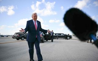 Een man in een donker pak met rode stropdas spreidt beide armen, terwijl hij over een asfaltweg loopt.