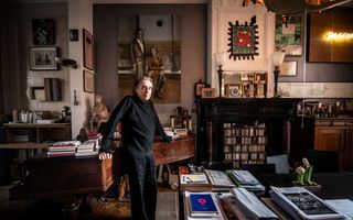 Oudere man met grijs achterovergekamd haar en zwarte kleding leunt tegen een met stapels boeken bedekte vleugel. Aan de muur hangen uiteenlopende kunstwerken.