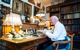 Een oudere man in een wit overhemd achter een bureau, met om zich heen veel kunstvoorwerpen.